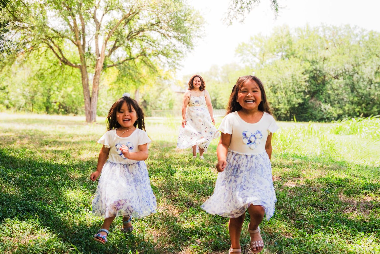 Three young girls in white dresses running and laughing through a sunny green park surrounded by trees.