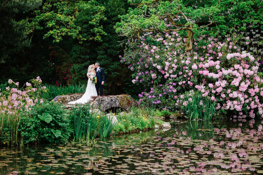 Bride and groom share a romantic moment by a serene pond surrounded by lush greenery and blooming pink flowers.