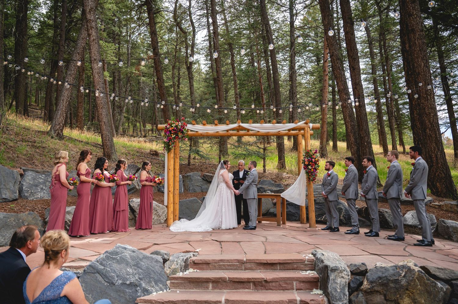 Outdoor wedding ceremony in forest with bride, groom, bridesmaids in burgundy, groomsmen under wooden arbor with guests seated.