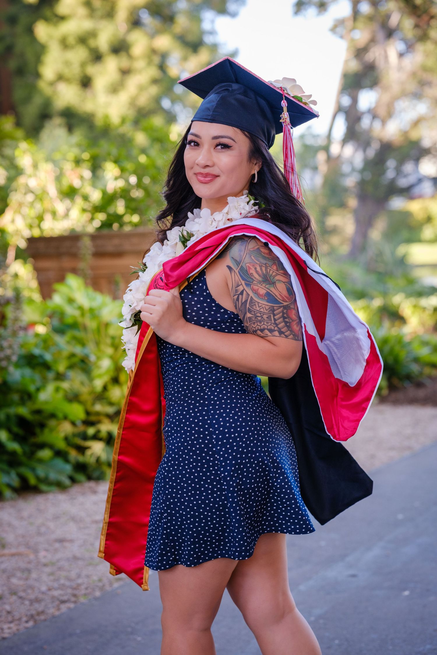 Smiling graduate in cap, gown, and floral lei poses joyfully on a tree-lined path, one leg raised in celebration.