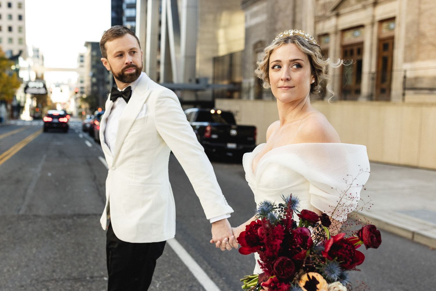 Bride and groom holding hands on a city street, groom in white tuxedo jacket.