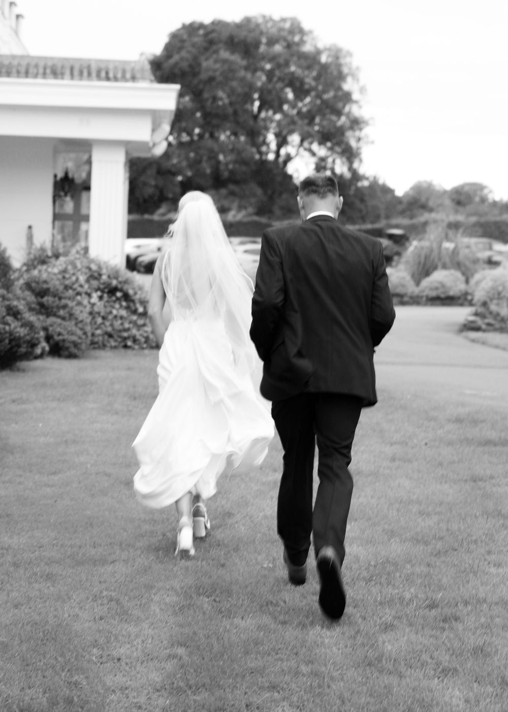 Black and white photo of bride and groom walking hand in hand across lawn toward elegant venue.