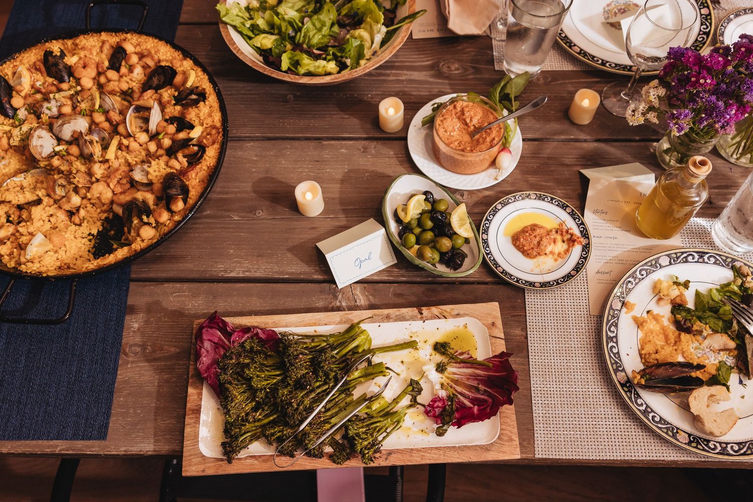 Overhead view of a rustic wooden table with Mediterranean dishes including paella, hummus, roasted asparagus, and fresh salads.
