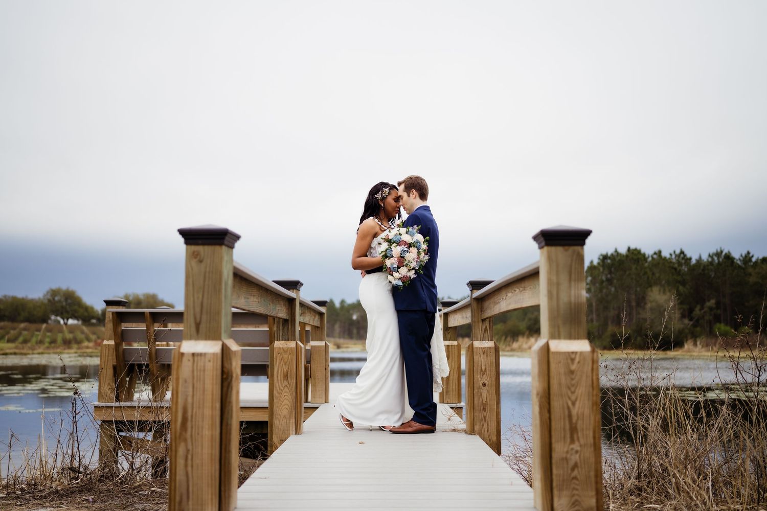 Wedding party members gather outdoors wearing burgundy dresses and navy suits while smiling and holding bouquets.