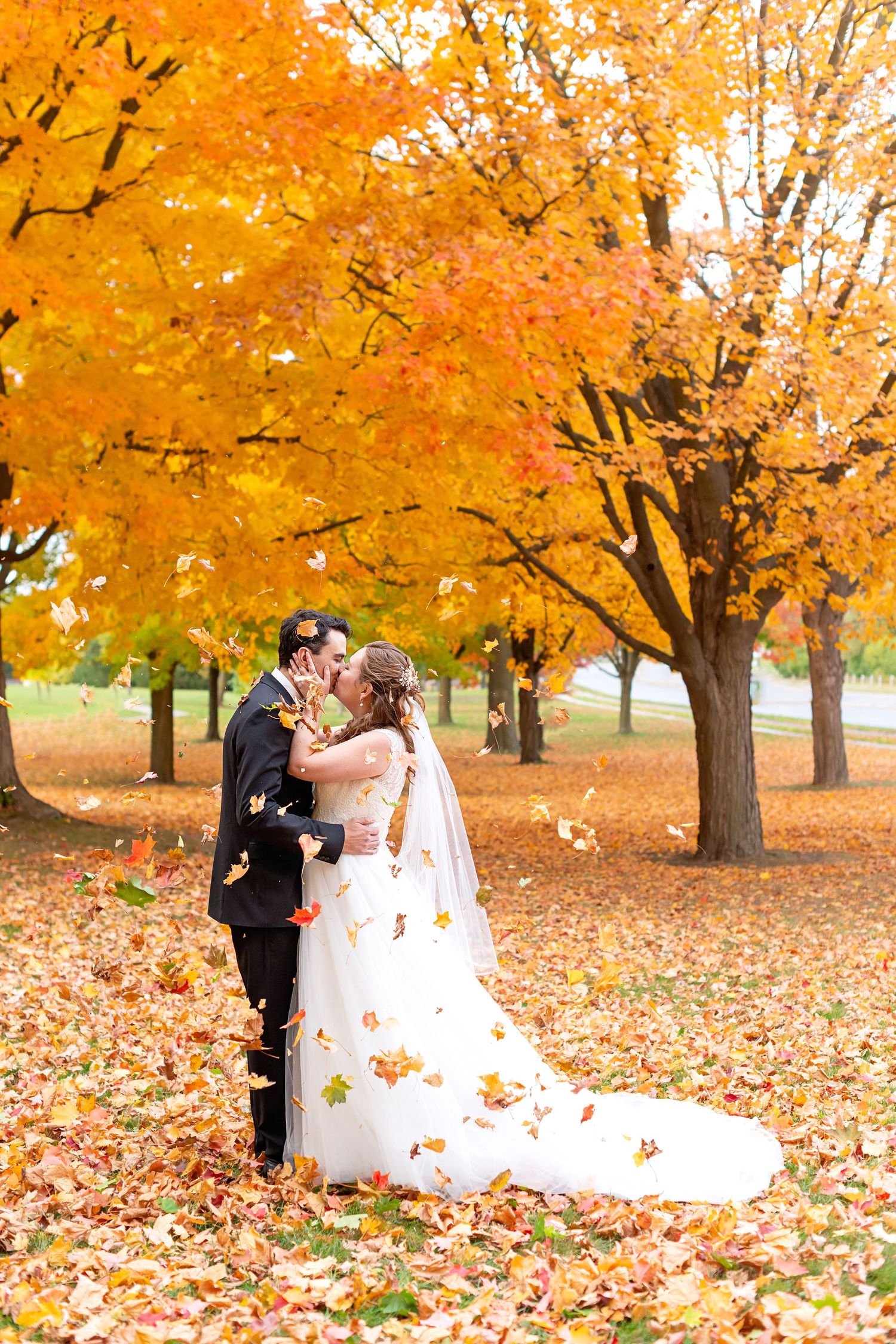 A romantic wedding couple embraces beneath vibrant orange autumn trees with fallen leaves scattered on the ground.
