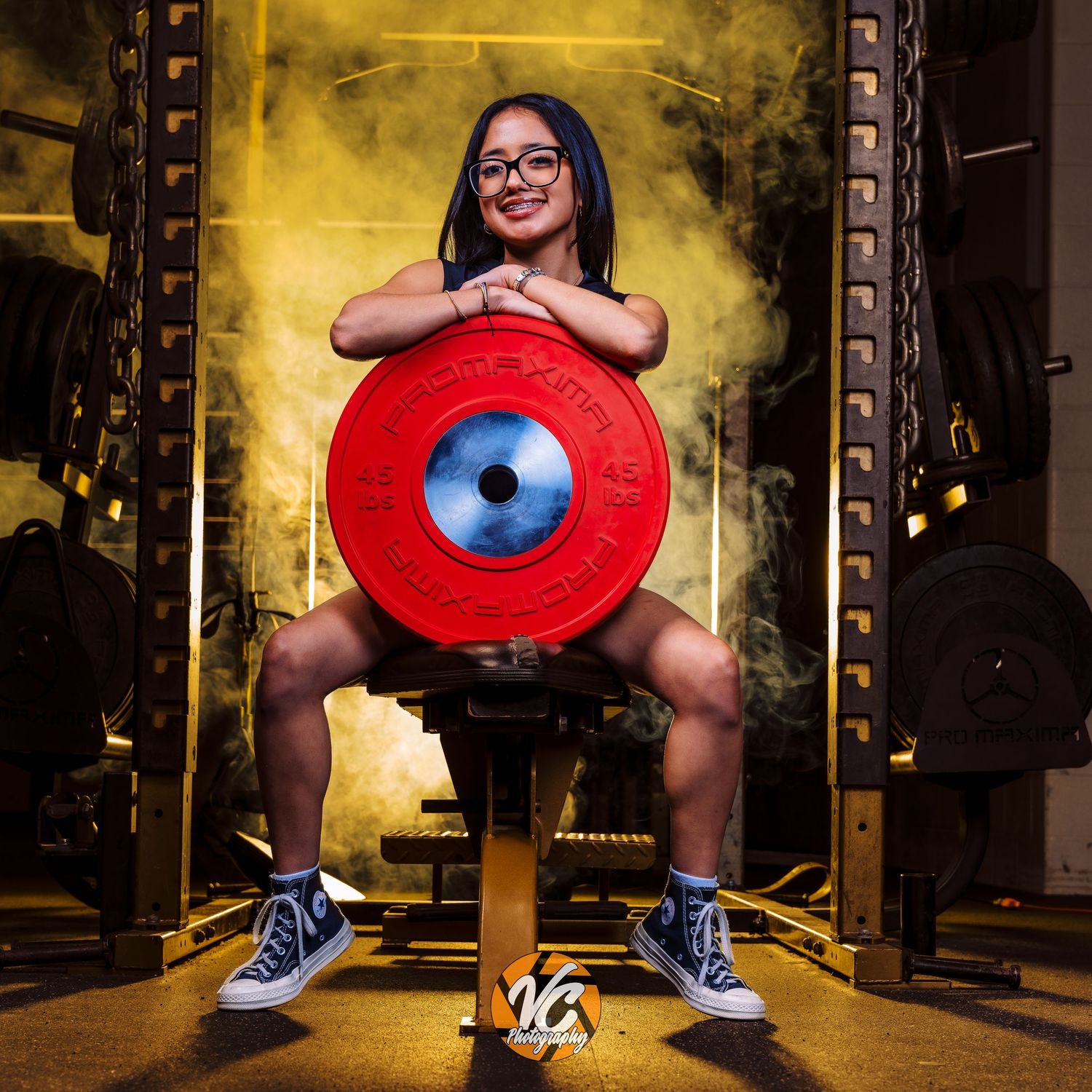 Young woman in glasses posing confidently in a gym holding a red bumper plate weight, wearing blue Converse sneakers.