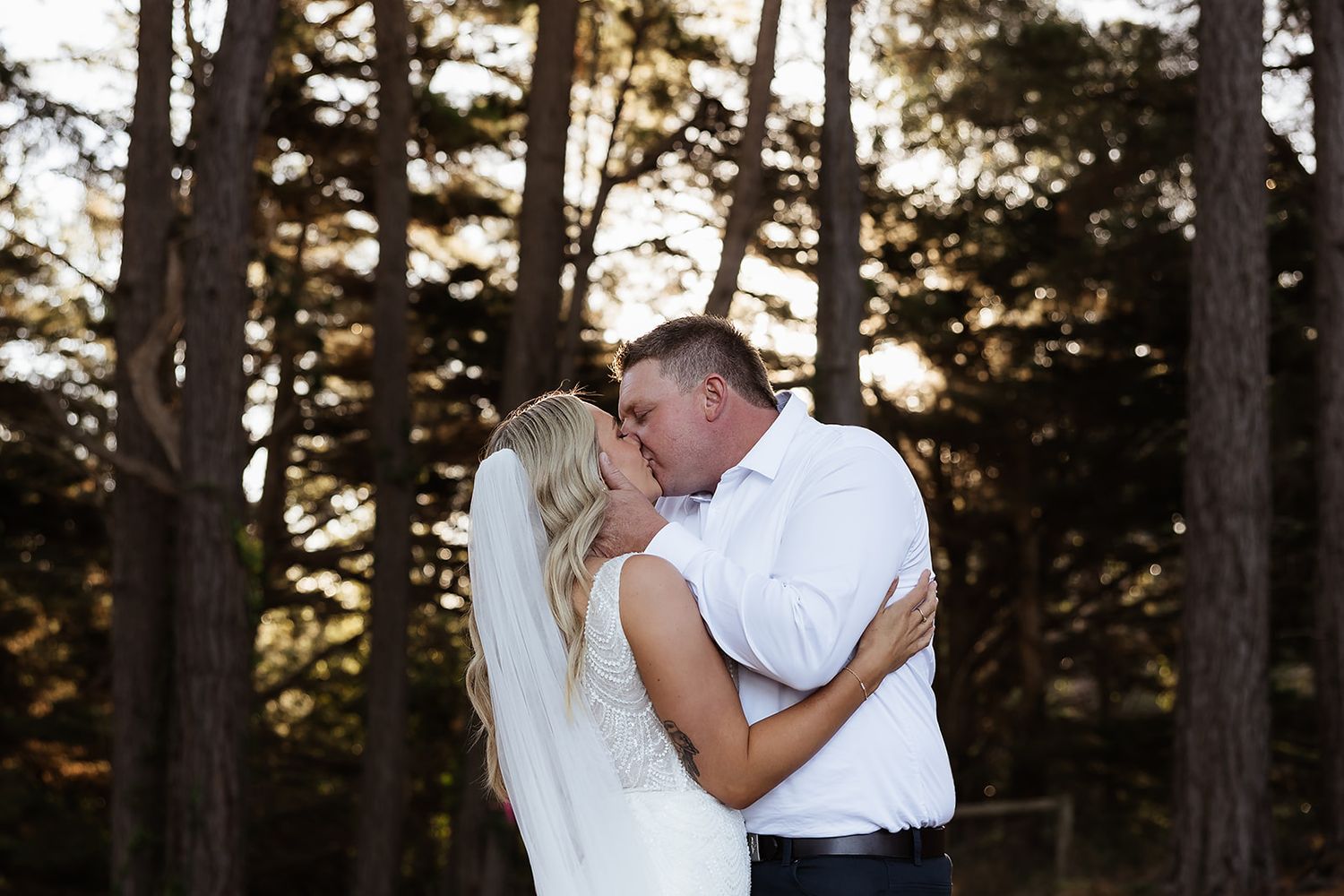 A bride and groom share a romantic kiss outdoors surrounded by tall pine trees in golden sunset light.