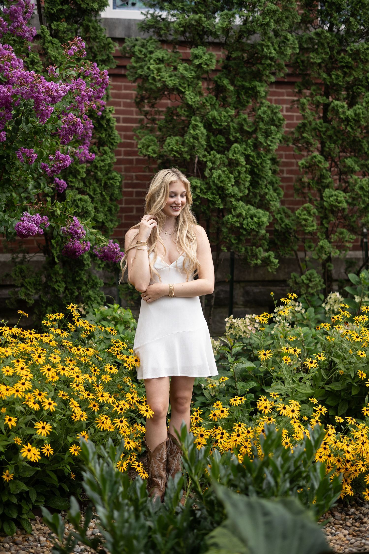 Smiling blonde woman in white dress and cowboy boots standing among yellow black-eyed Susans and purple flowers.