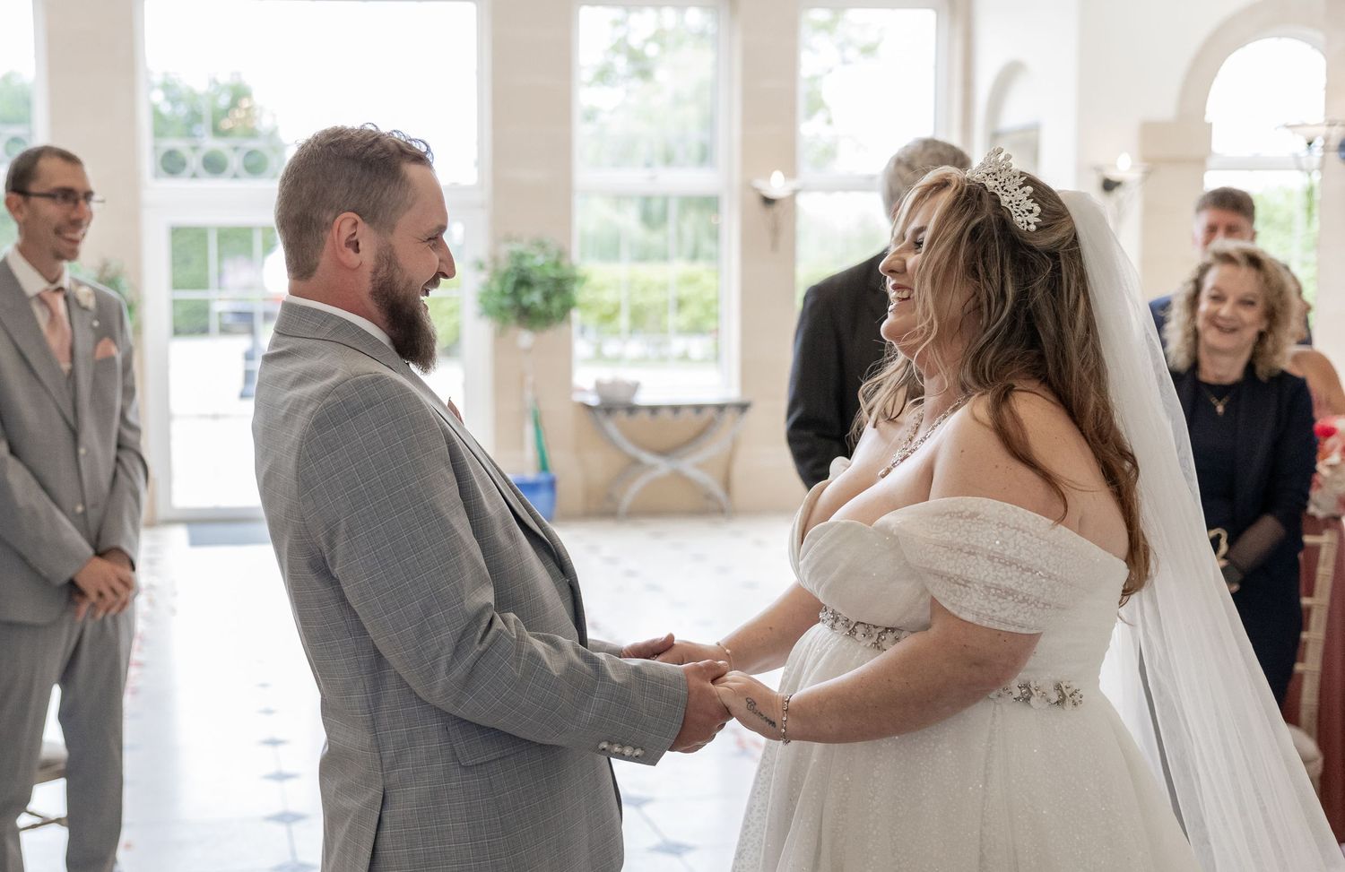 Bride and groom share a romantic moment by a large tree, bride in white off-shoulder gown with veil, groom in gray suit.