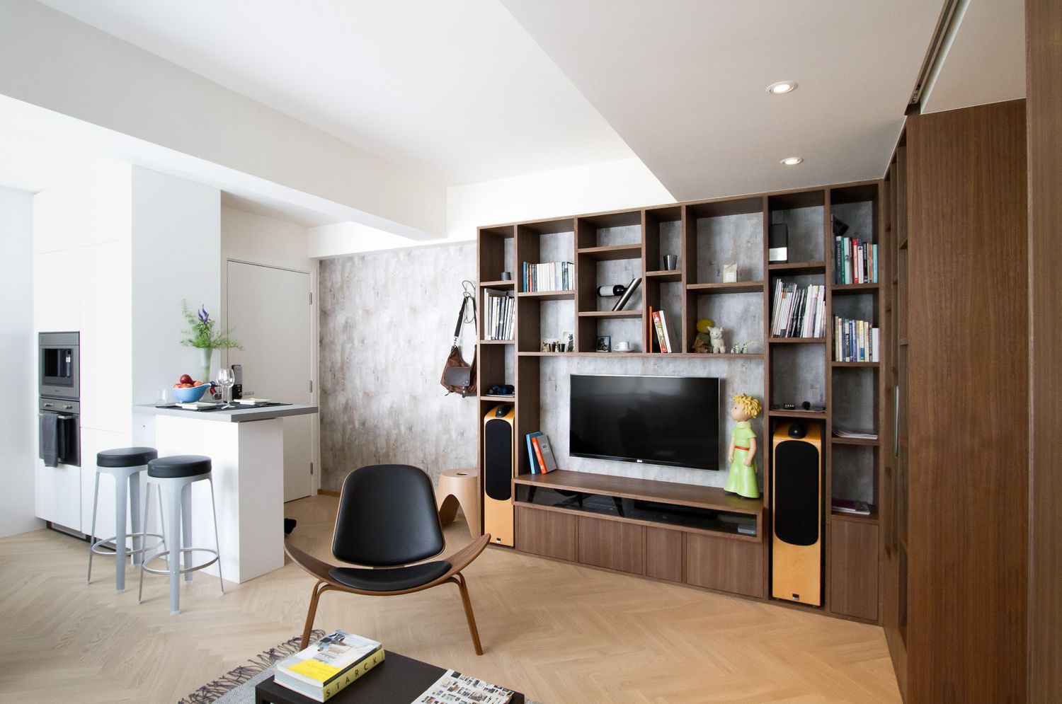 Modern living room with walnut shelving unit, flat screen TV, designer chair, and open kitchen.