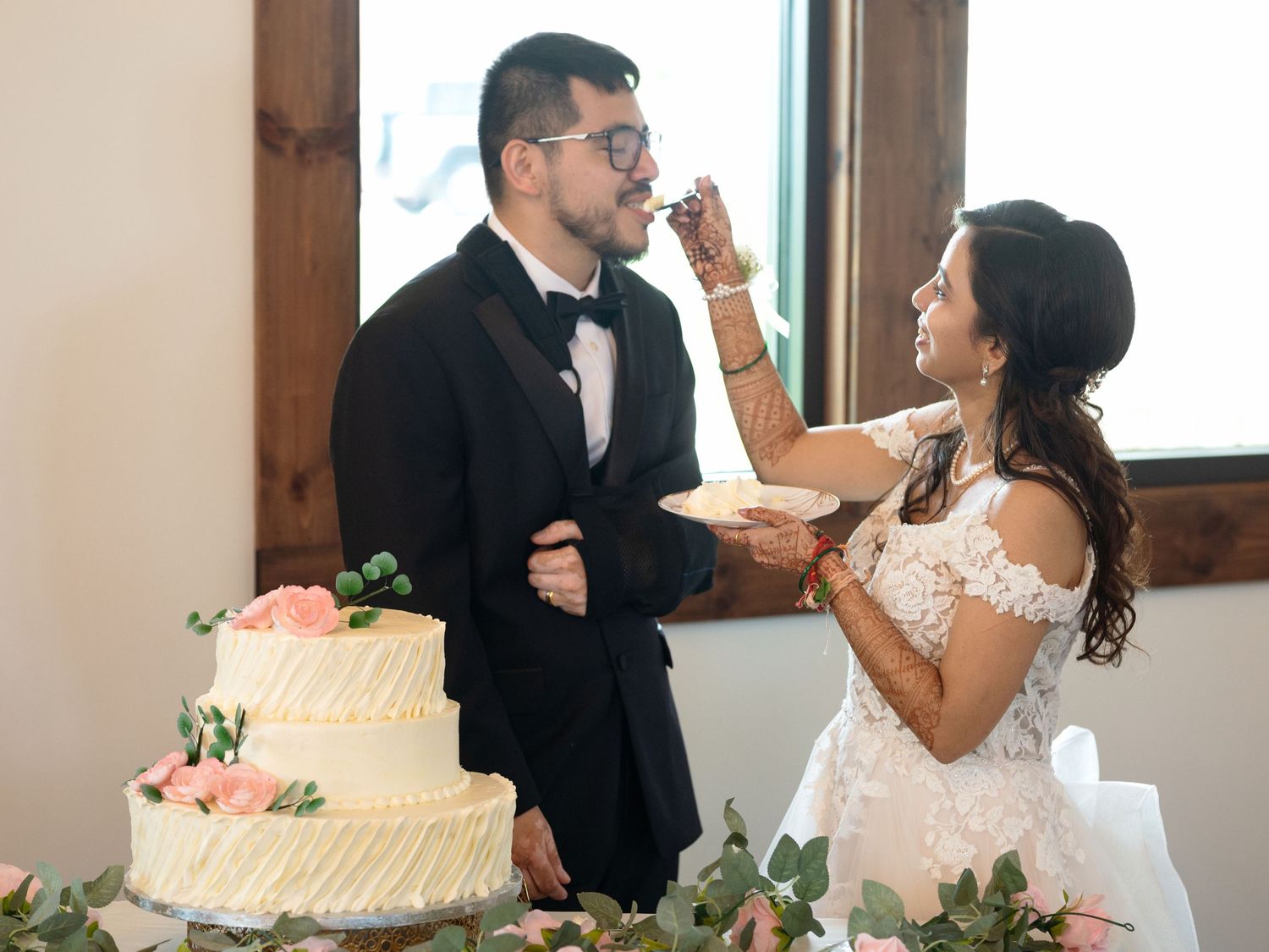 Bride feeds groom wedding cake at reception, three-tier white cake with pink roses on table nearby.