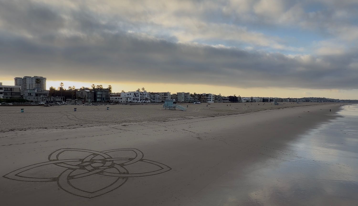 Serene beach at dusk with geometric sand art, calm shoreline, and coastal buildings under dramatic cloudy sky.