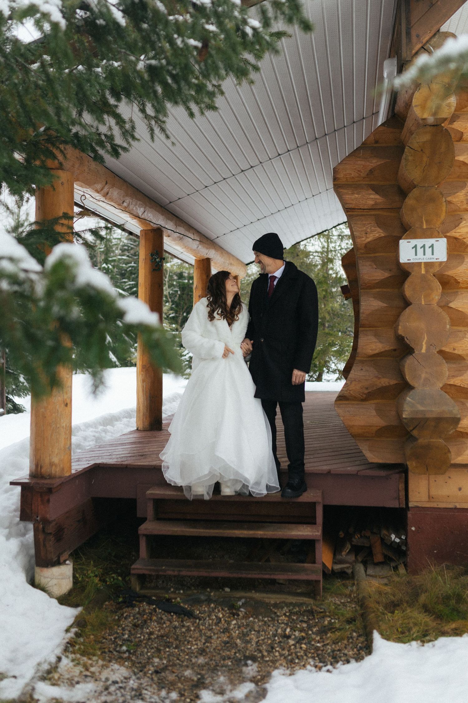 bride and groom walk along porch of cedar log cabin in the snowy trees during Revelstoke elopement