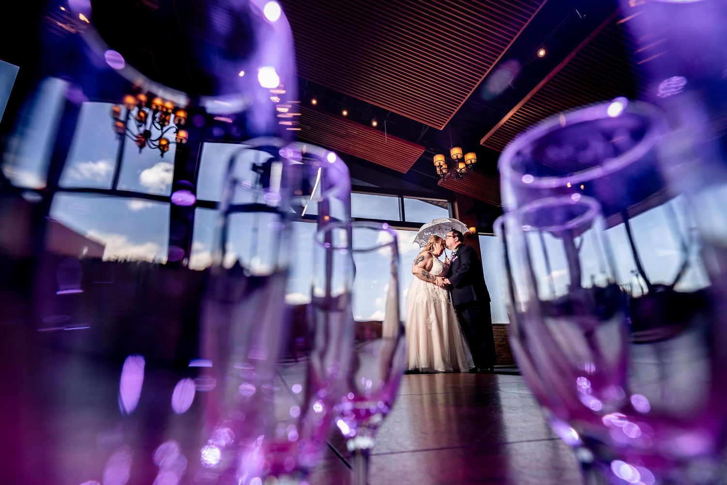 Bride and groom embracing in elegant wedding venue with purple lighting and champagne glasses in foreground.
