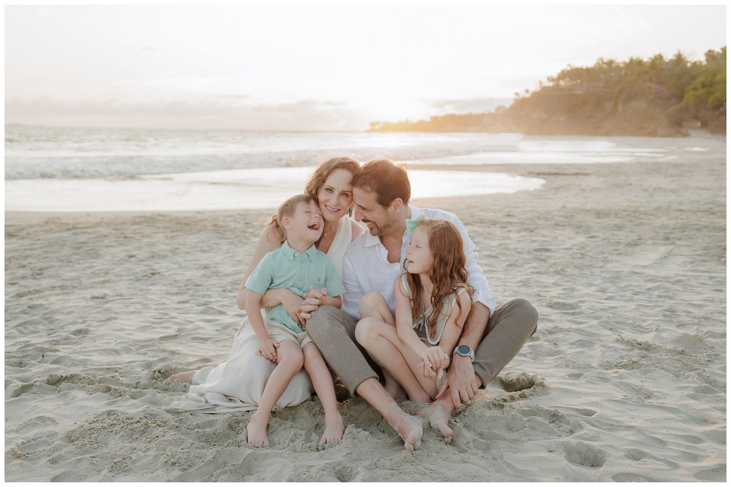 A family plays together on a beach, lifting a child in the air while wearing white clothing in black and white photography.