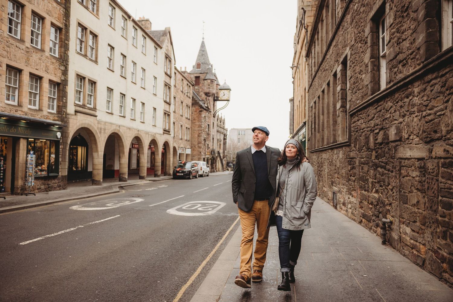 Couple walking hand in hand on a historic cobblestone street in Edinburgh, Scotland, surrounded by stone buildings.