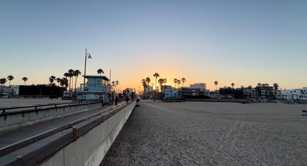 Golden sunset over Venice Beach boardwalk with palm trees, sandy shore, and lifeguard tower in Los Angeles, California.