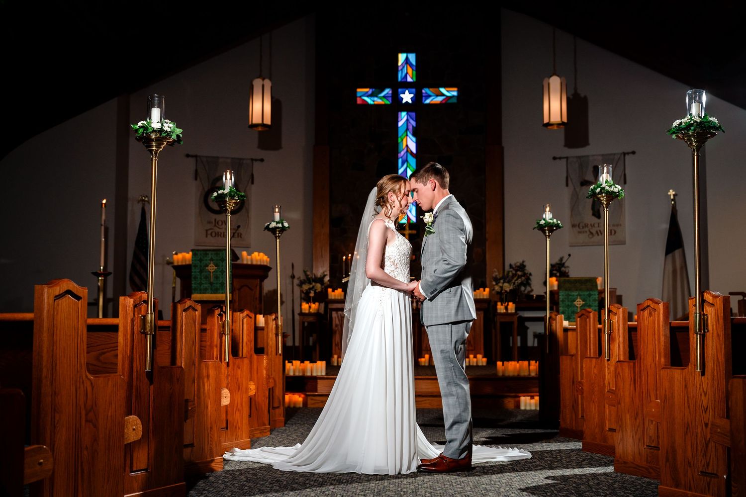 A couple shares a romantic kiss in a lush green garden, bride in white gown and groom in gray suit under blue sky.