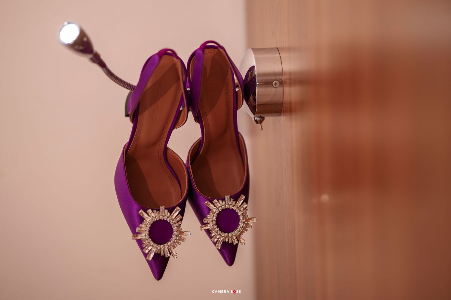 Purple pointed-toe heels with gold sunburst embellishments hanging on a door hook against a pink wall.