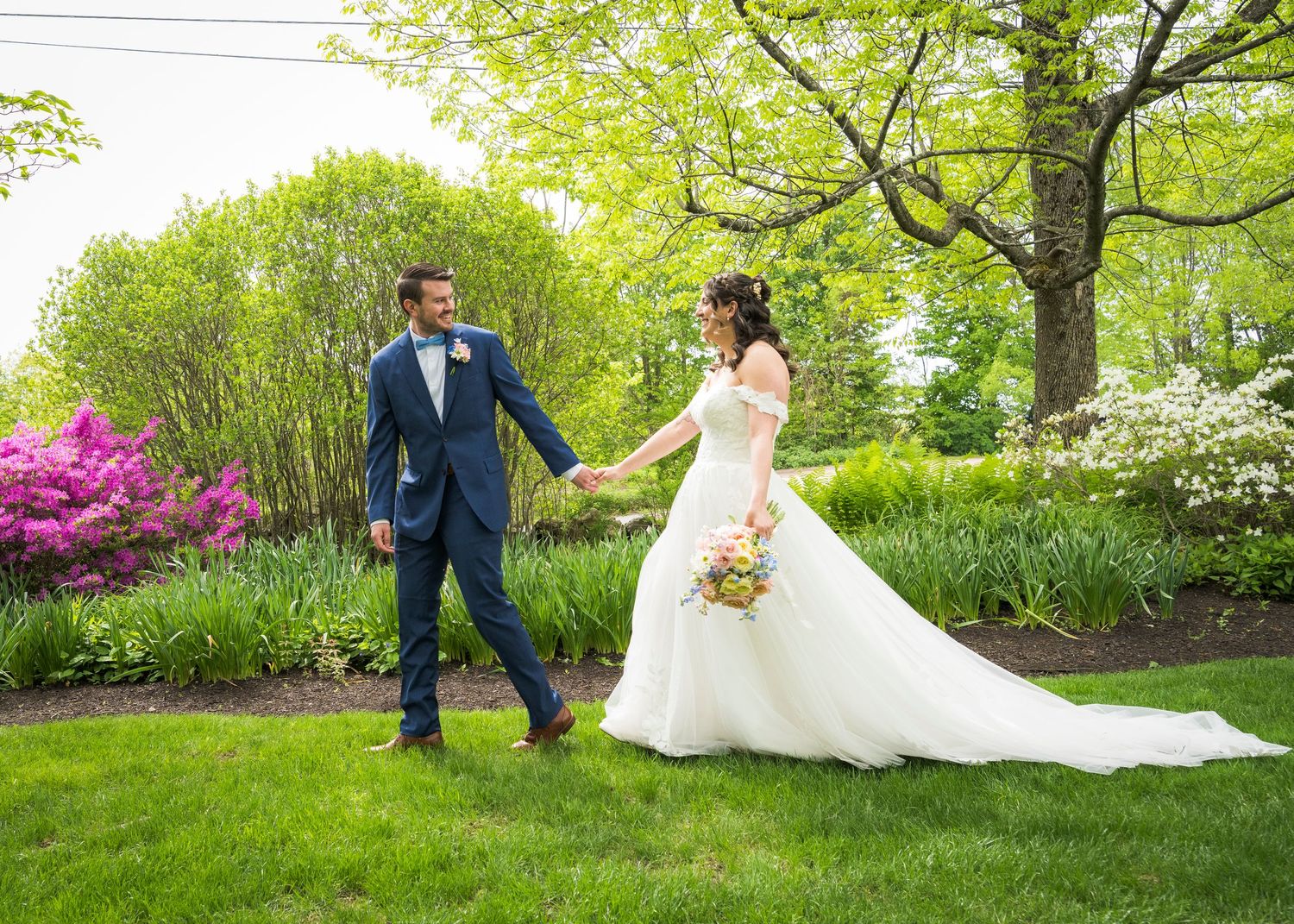Bride and groom walk hand-in-hand through a lush garden, surrounded by blooming flowers on their wedding day.