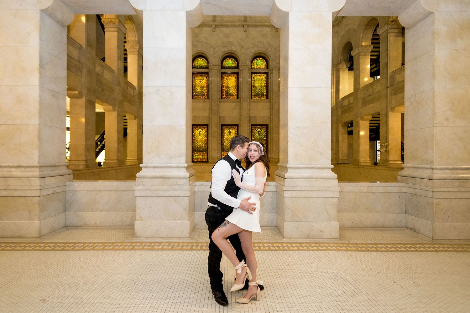 A couple embraces in a romantic moment inside an elegant building with arched windows and stone columns.