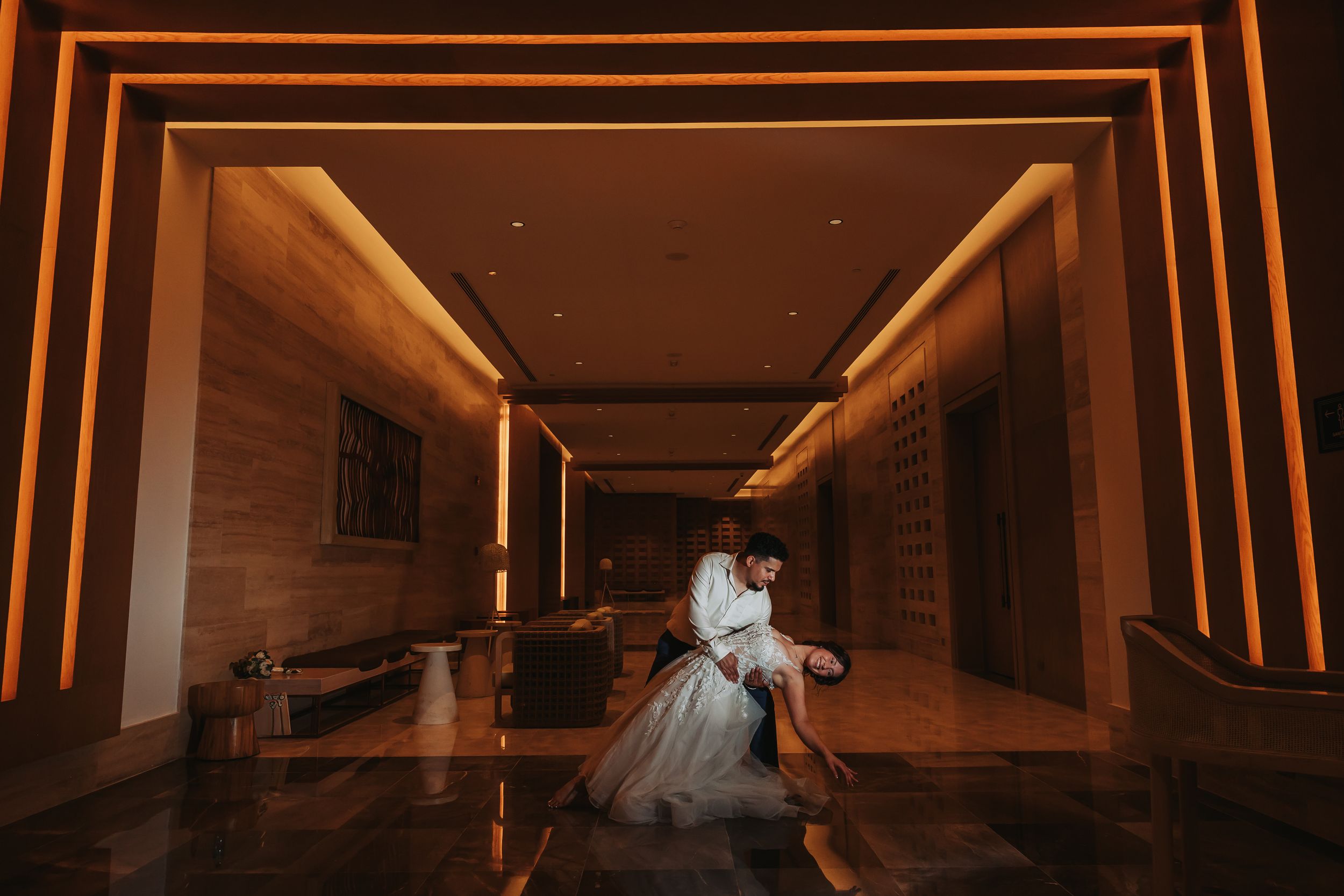 Couple sharing a romantic dip kiss in a warmly lit hotel lobby with glowing amber archways.