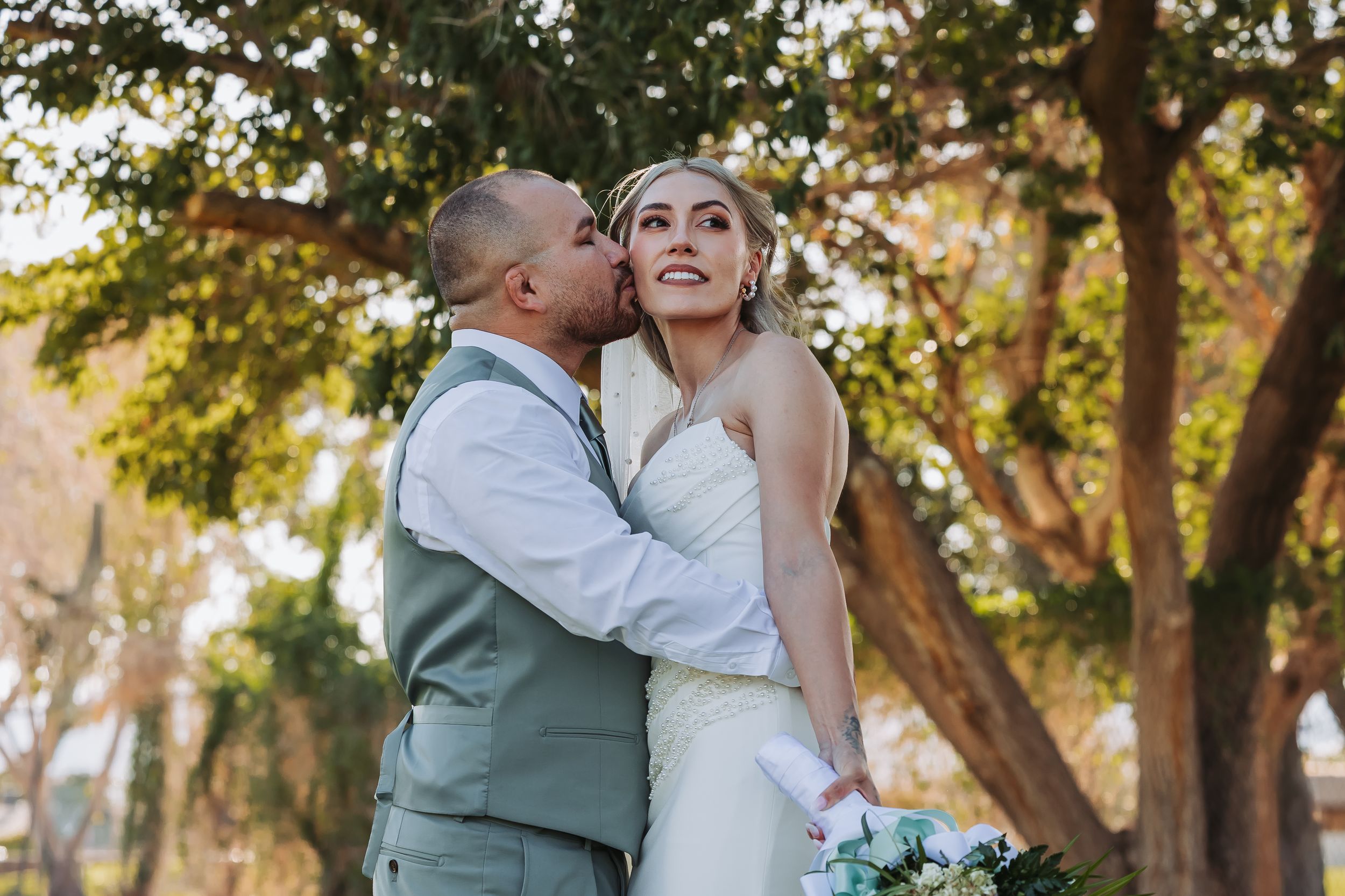 Groom in gray suit kissing bride on cheek outdoors under trees, bride smiling holding white bouquet.