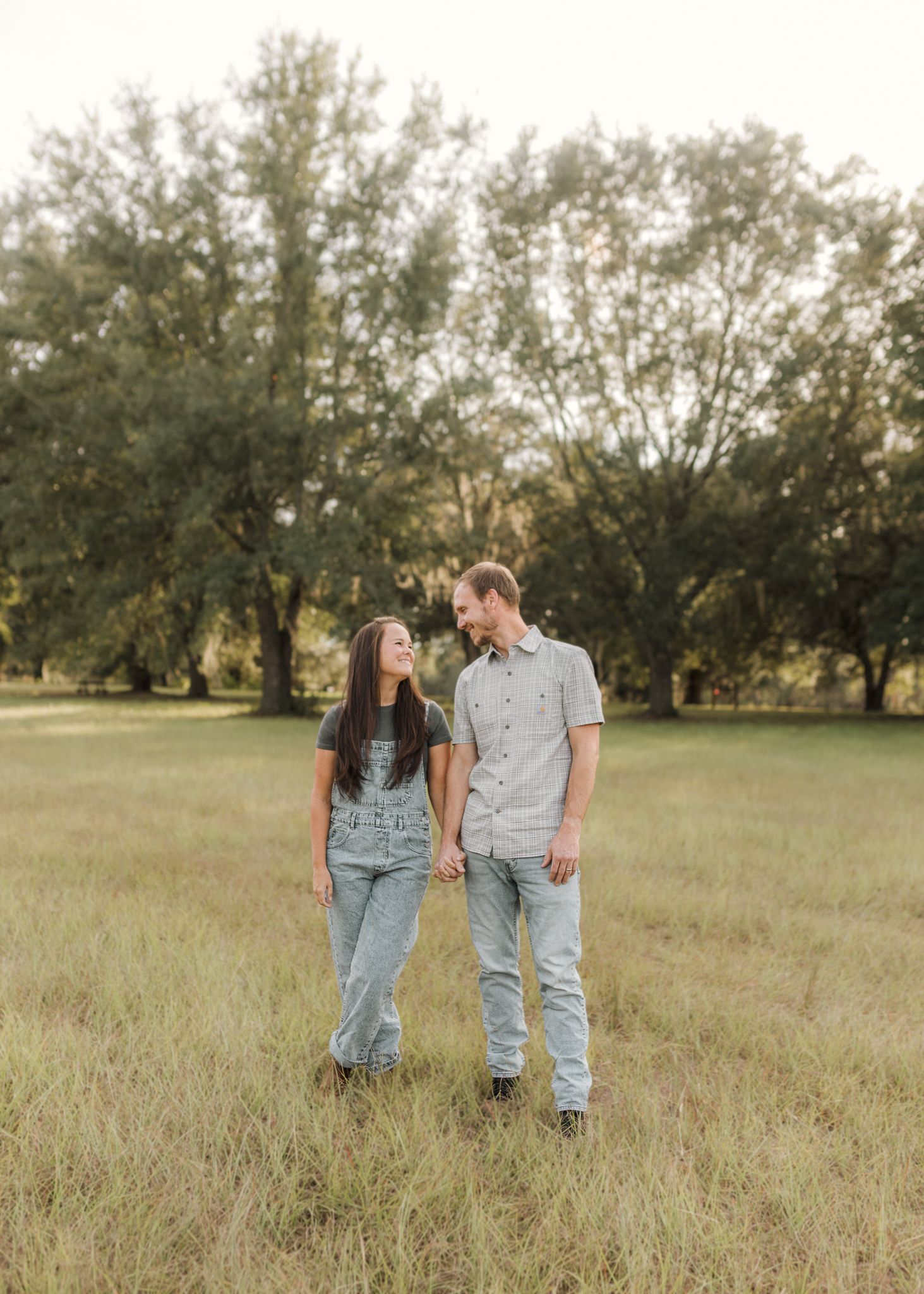 A couple holds hands and smiles at each other in a lush green field with trees in the background.