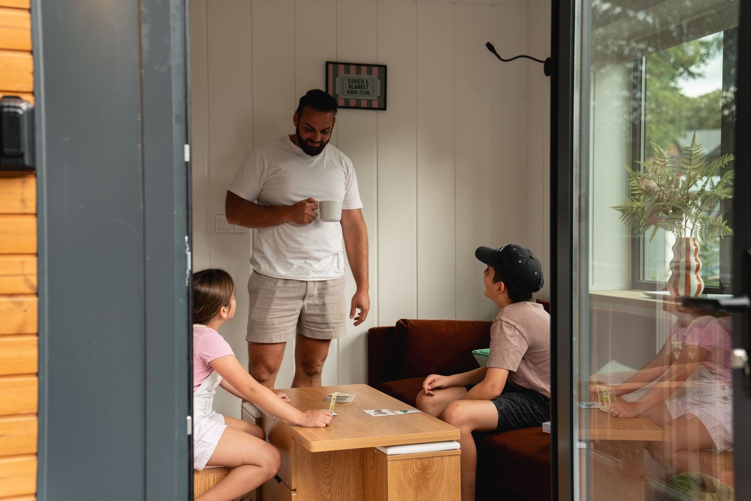 A man stands talking with children around a wooden coffee table in a cozy outdoor cabin porch seating area.