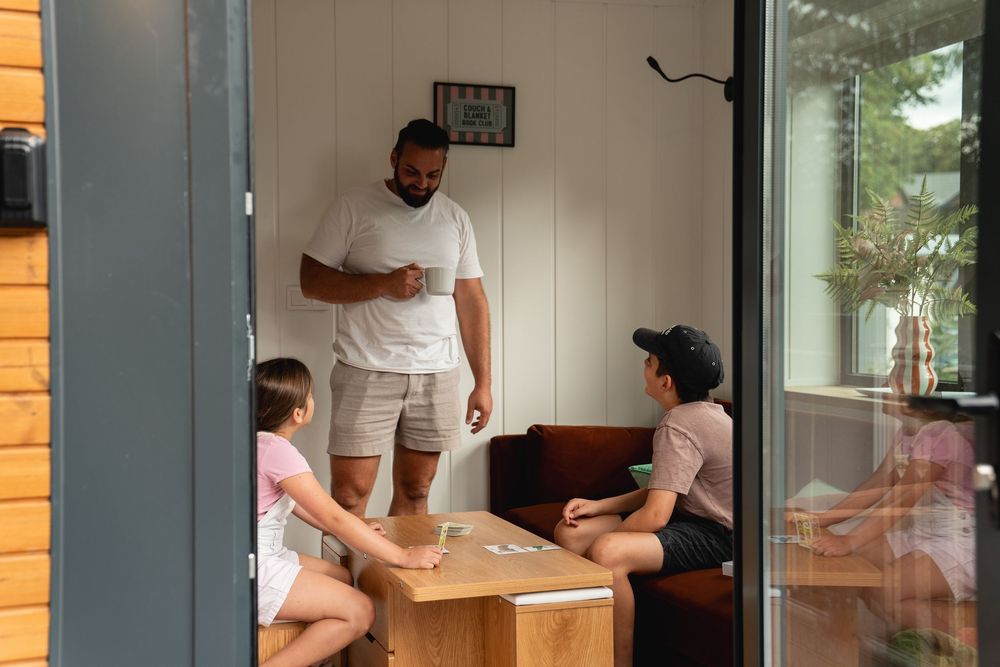 A man stands talking with children around a wooden coffee table in a cozy outdoor cabin porch seating area.