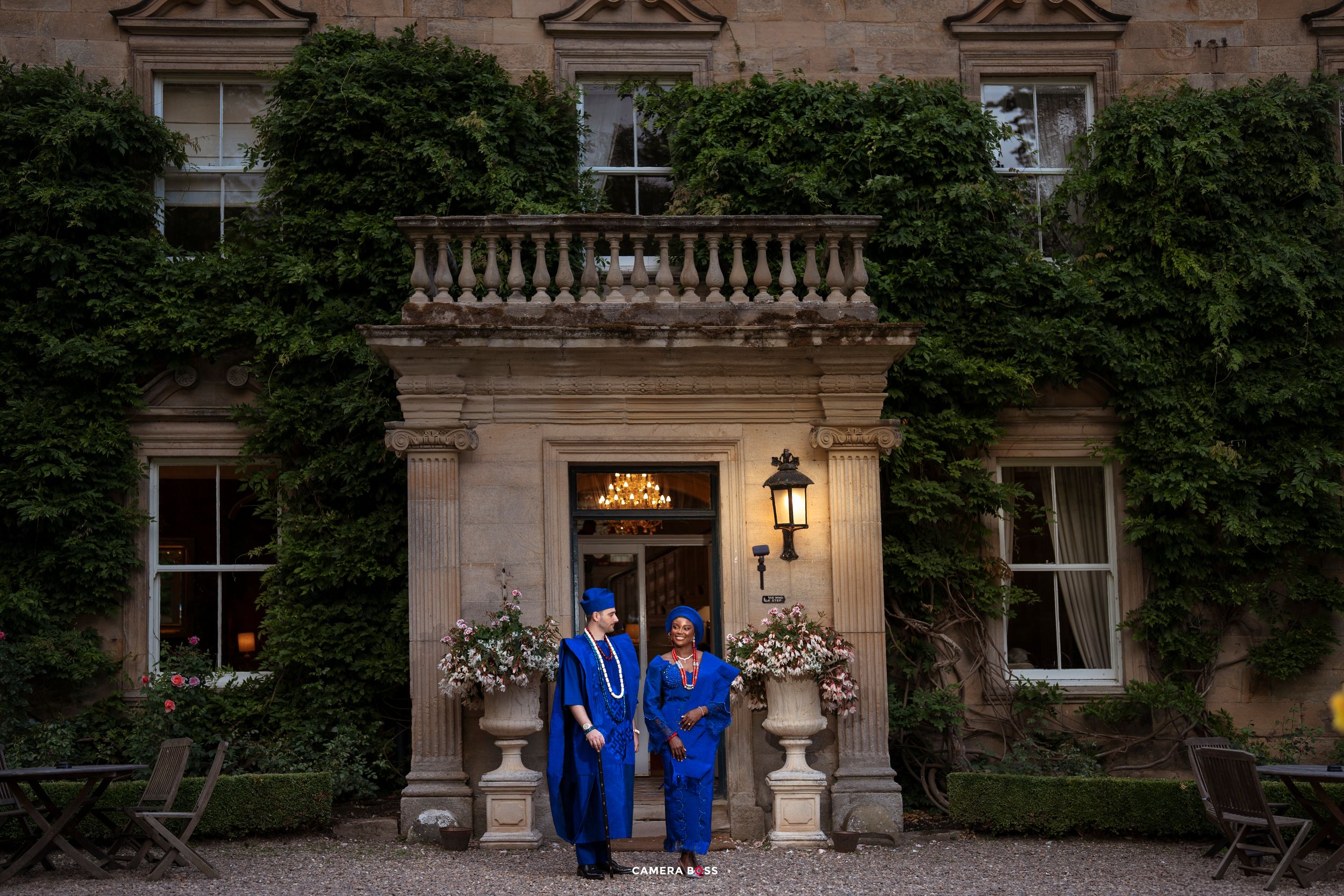 Two graduates in blue robes and caps pose outside an ivy-covered historic mansion at dusk.