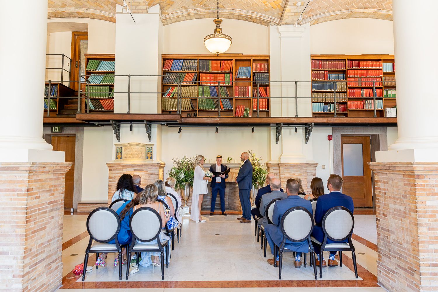 Intimate wedding ceremony in a stunning library venue with colorful bookshelves and elegant blue chairs.