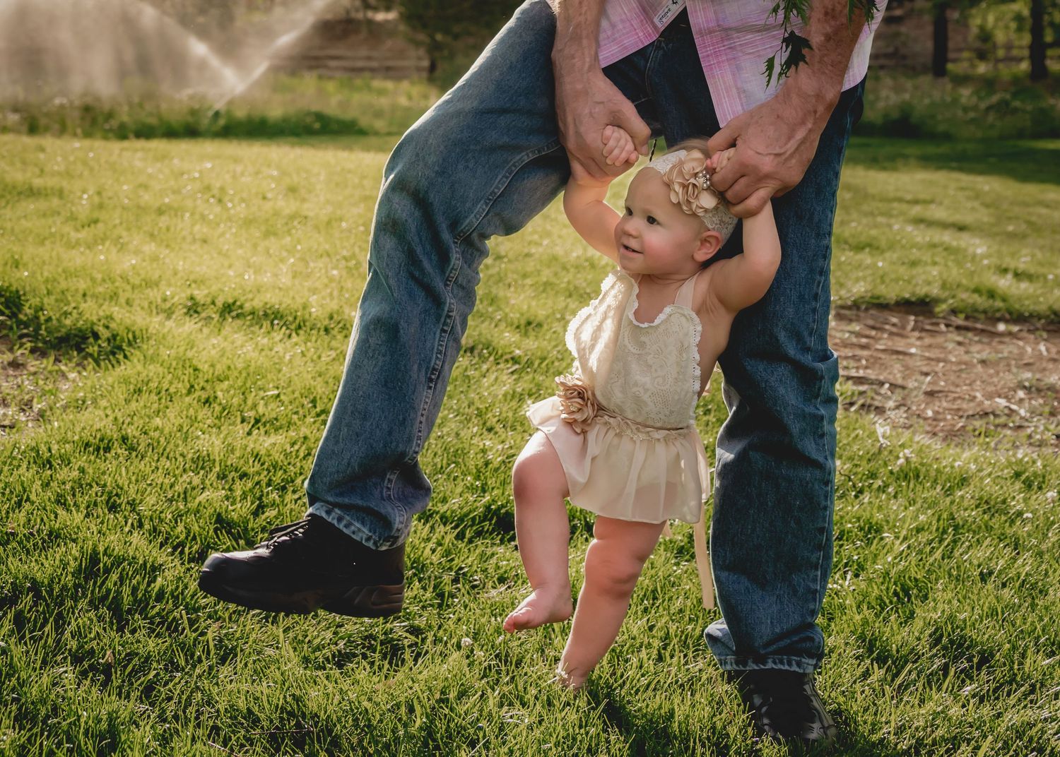 A toddler in a white romper takes first steps outdoors, supported by an adult holding her hands on green grass.