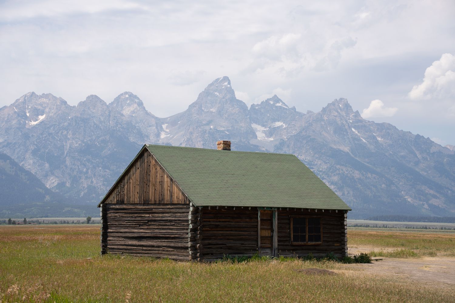 Historic log cabin with green roof sits in open meadow with snow-capped Grand Teton mountain range in background.