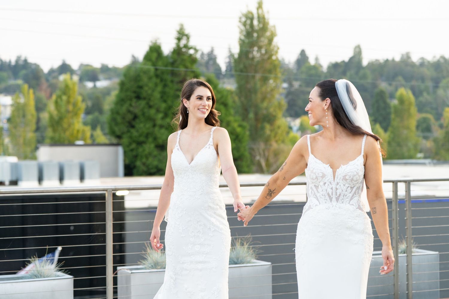 Two brides in white wedding gowns hold hands on a rooftop, smiling joyfully on their wedding day.