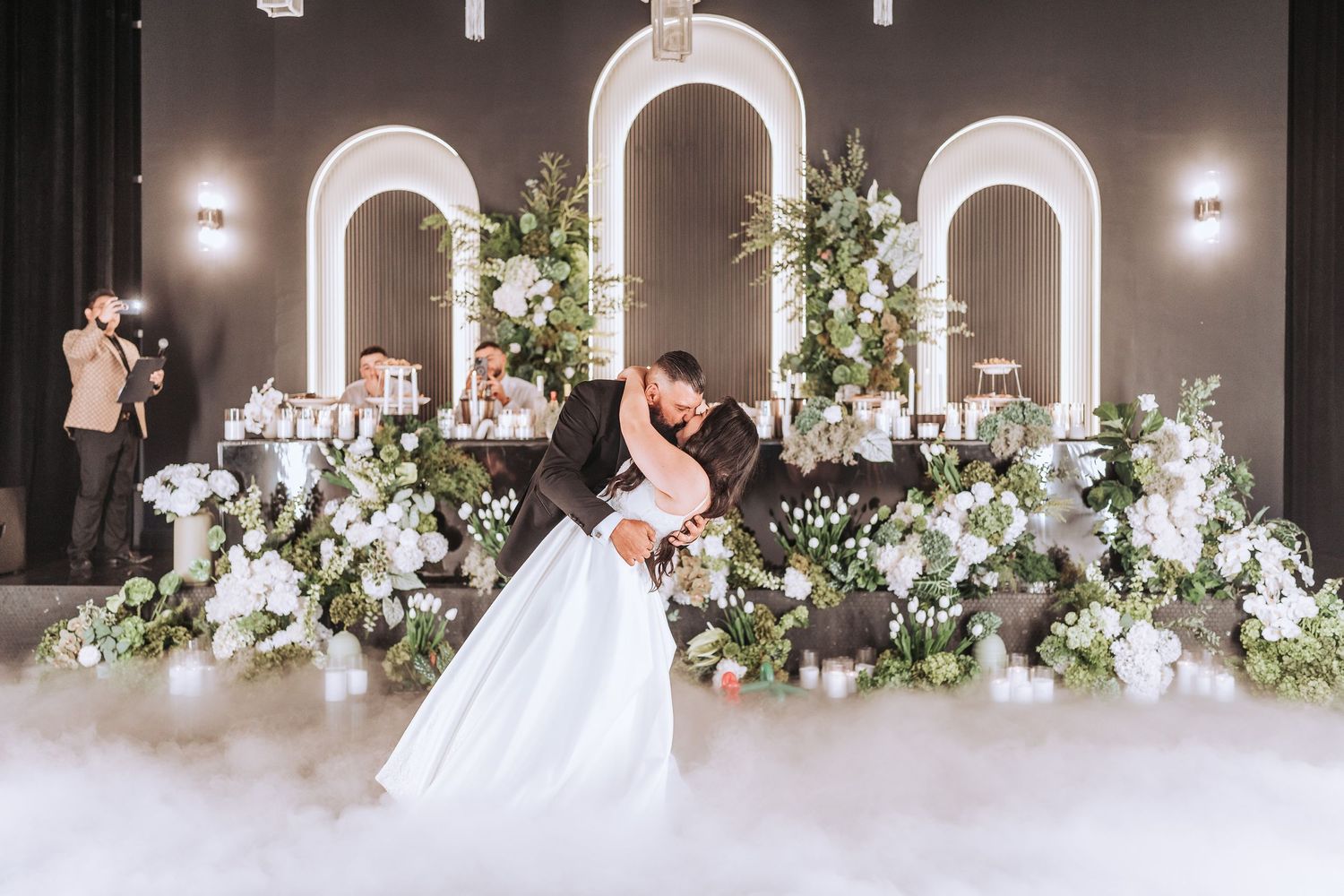 A couple dances in a romantic ballroom setting with white arched backdrops and elegant floral arrangements.
