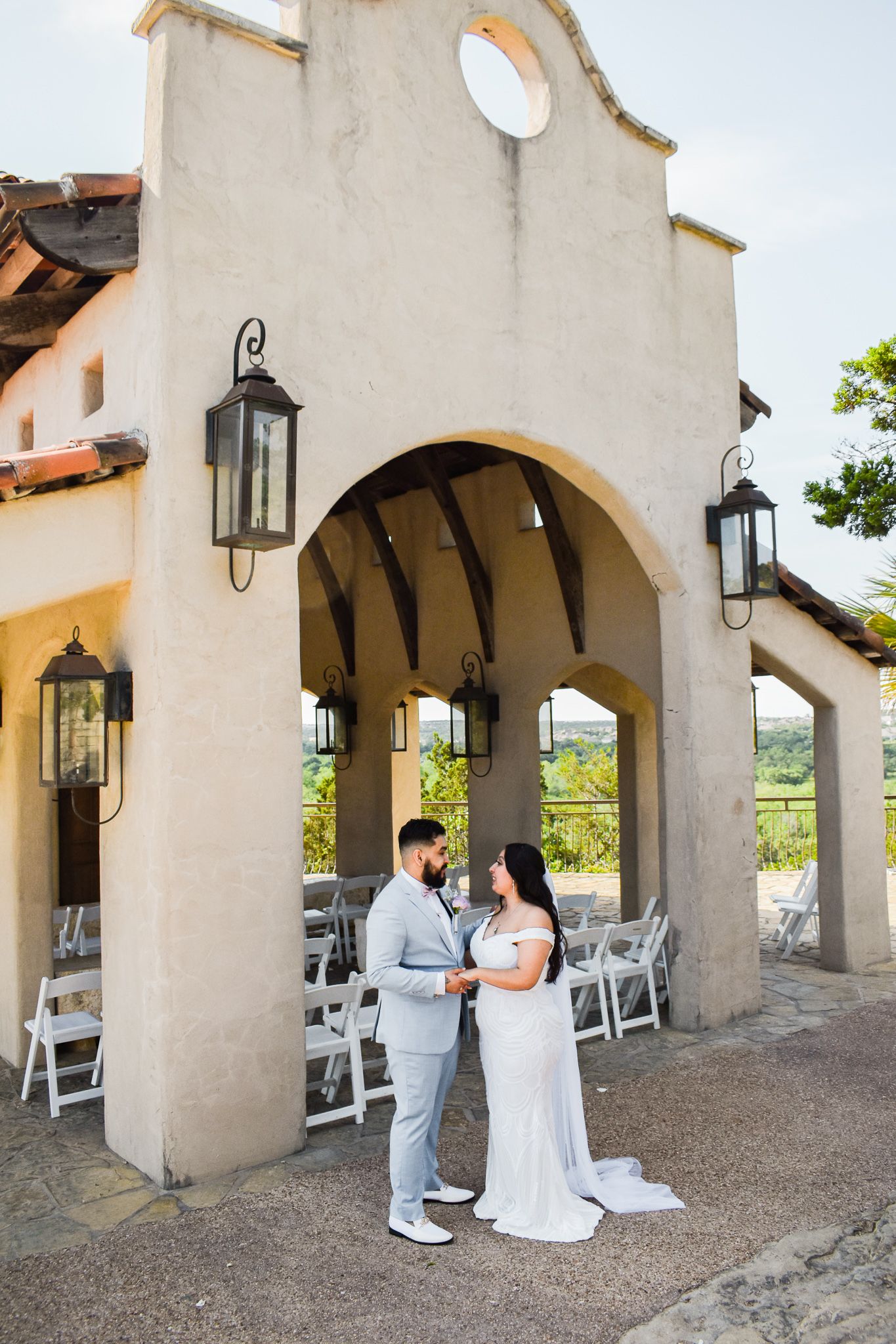 Bride and groom standing under Spanish archway with folding chairs in background.