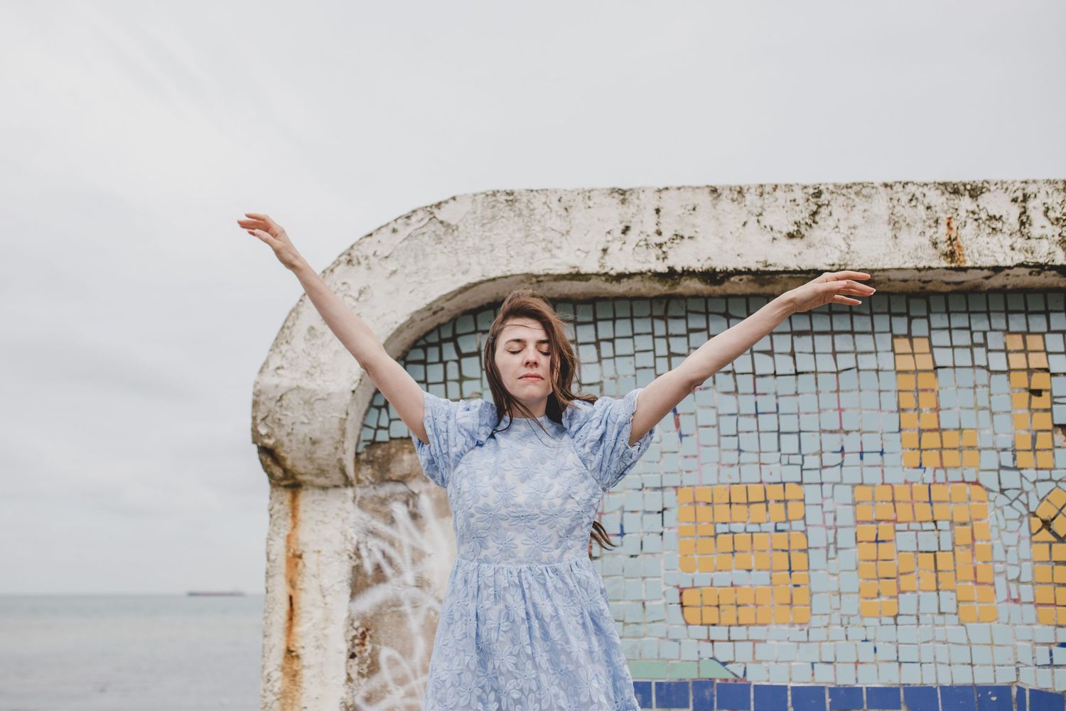 Person in blue dress celebrating with arms raised against vintage seaside wall.
