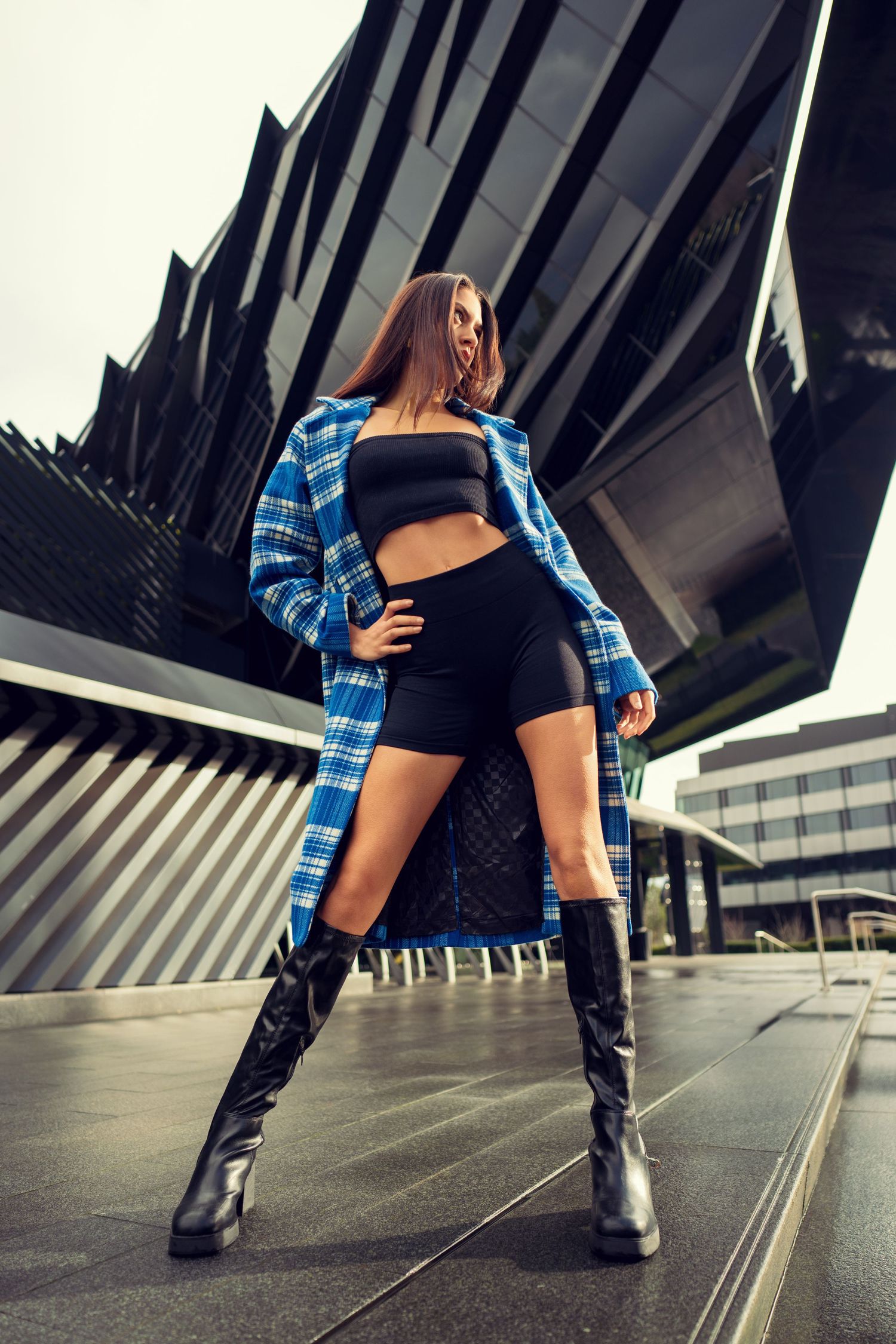 Woman in plaid blue flannel and black shorts poses in tall boots against modern architectural background.