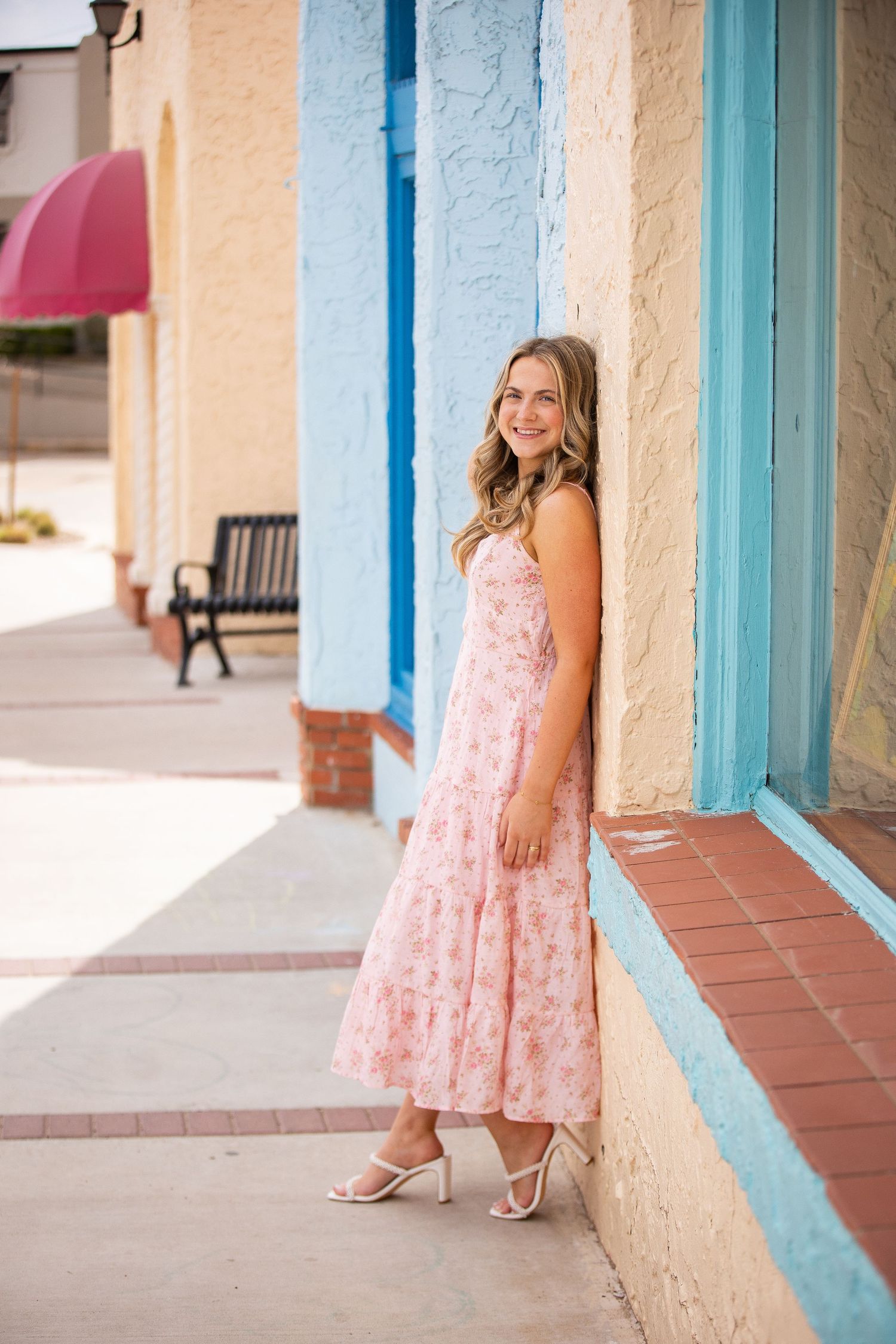 Woman in pink floral dress stands against colorful building facade with turquoise trim.