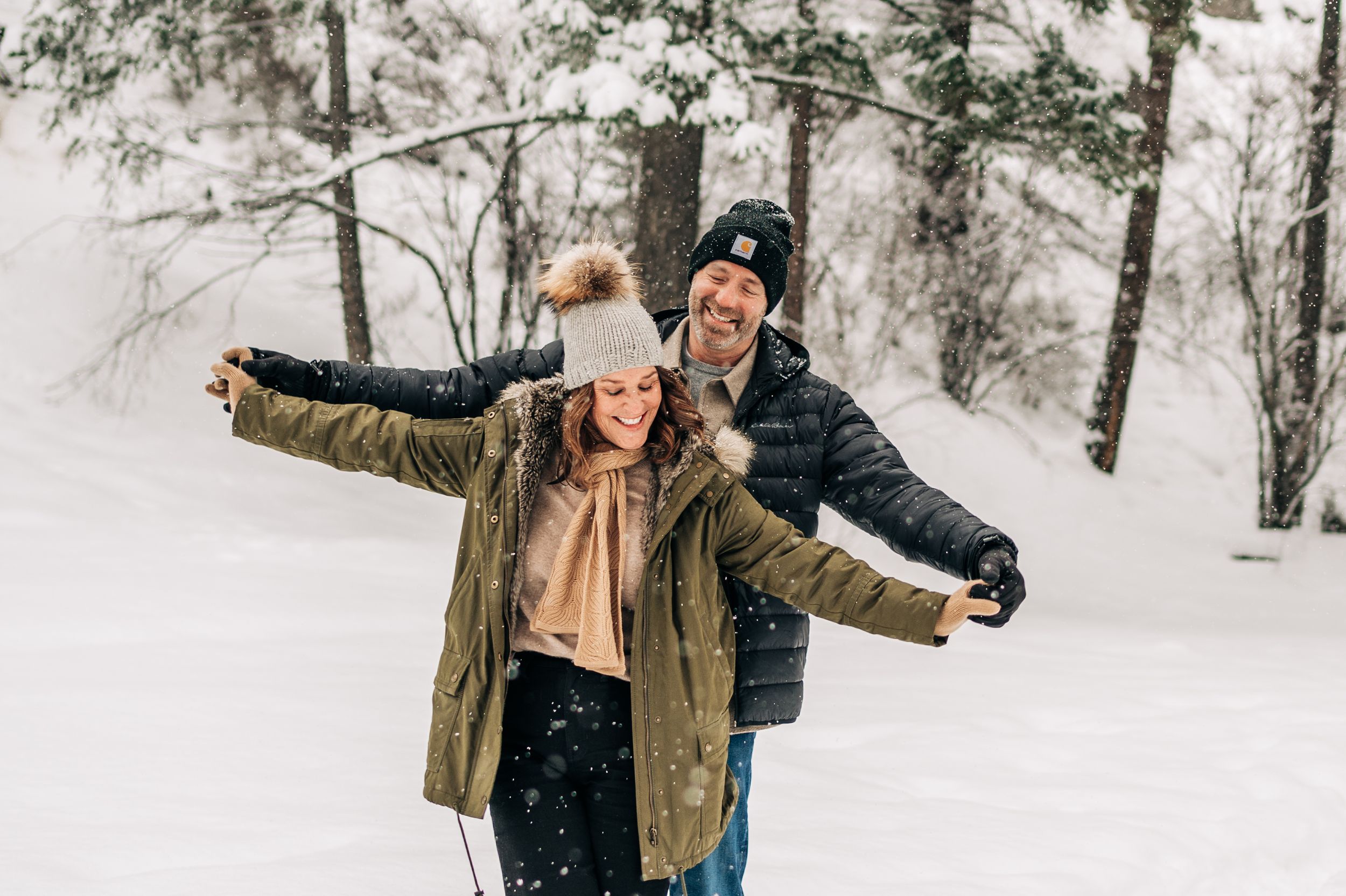 Two people in winter coats and knit hats spread their arms while enjoying a snowy forest scene together.
