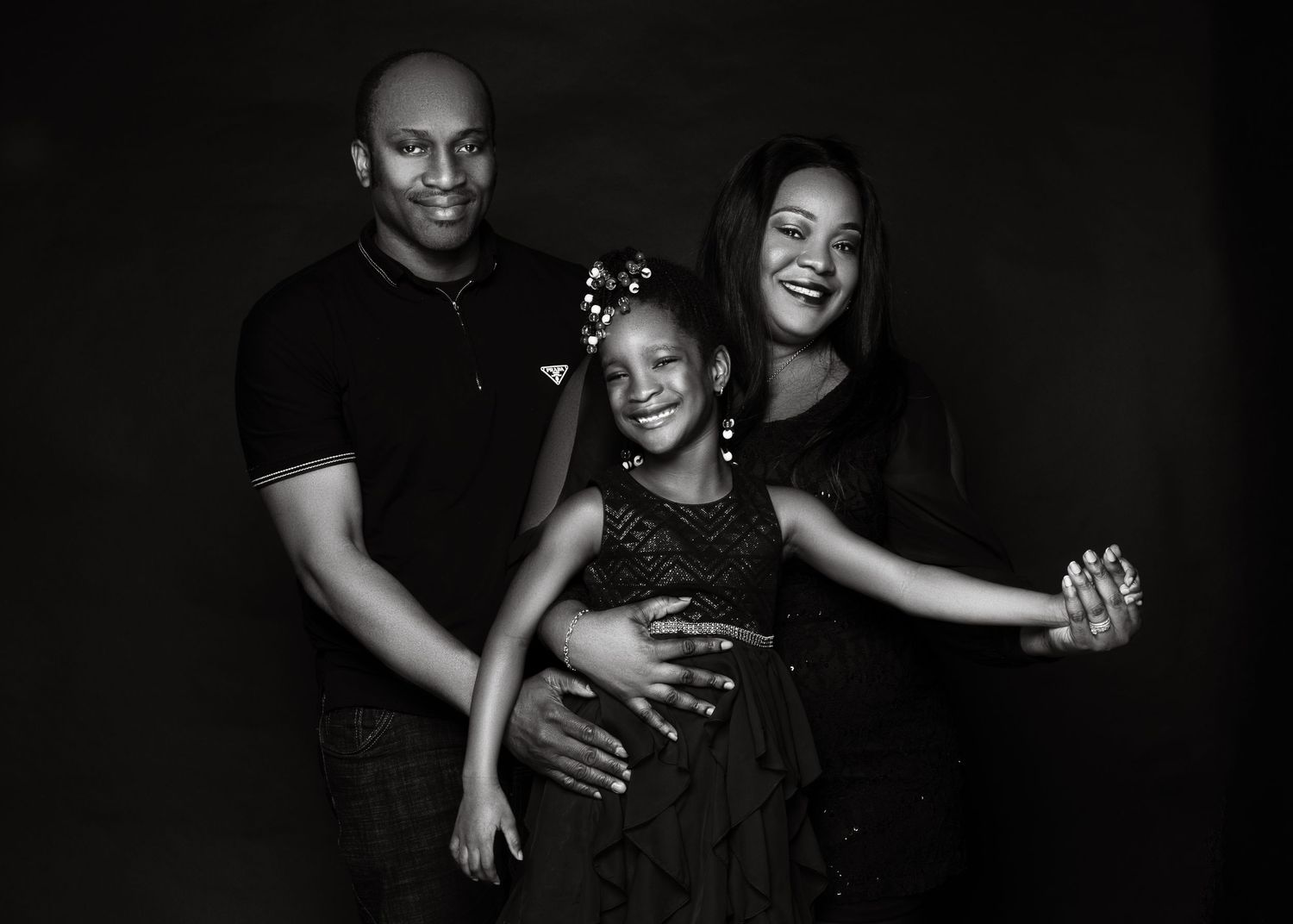 A black and white family portrait shows three members in a joyful embrace against a dark background.