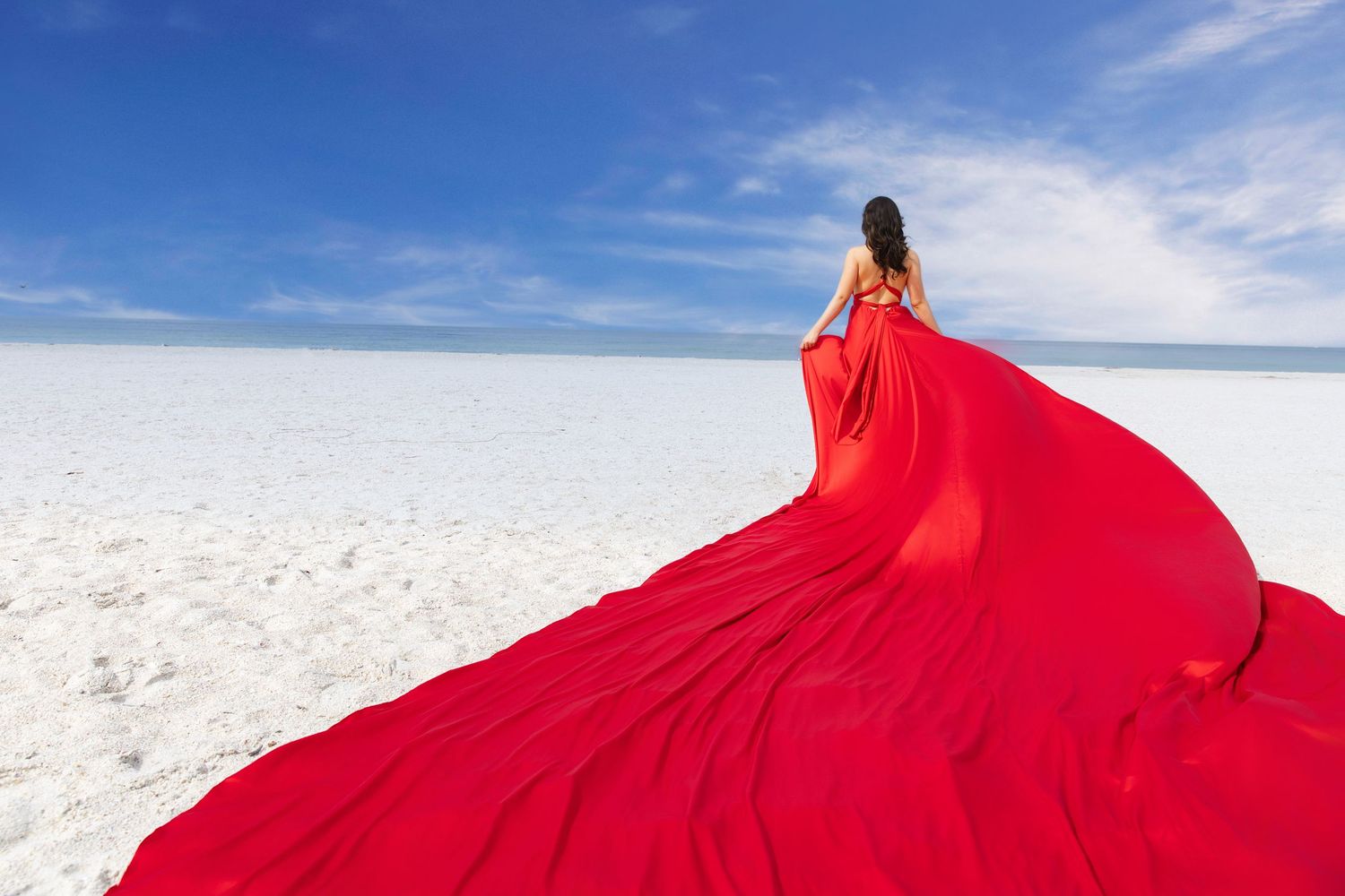 A flowing red dress billows dramatically against a scenic beach landscape with blue sky and clouds at sunset. Flying Dress Photoshoot