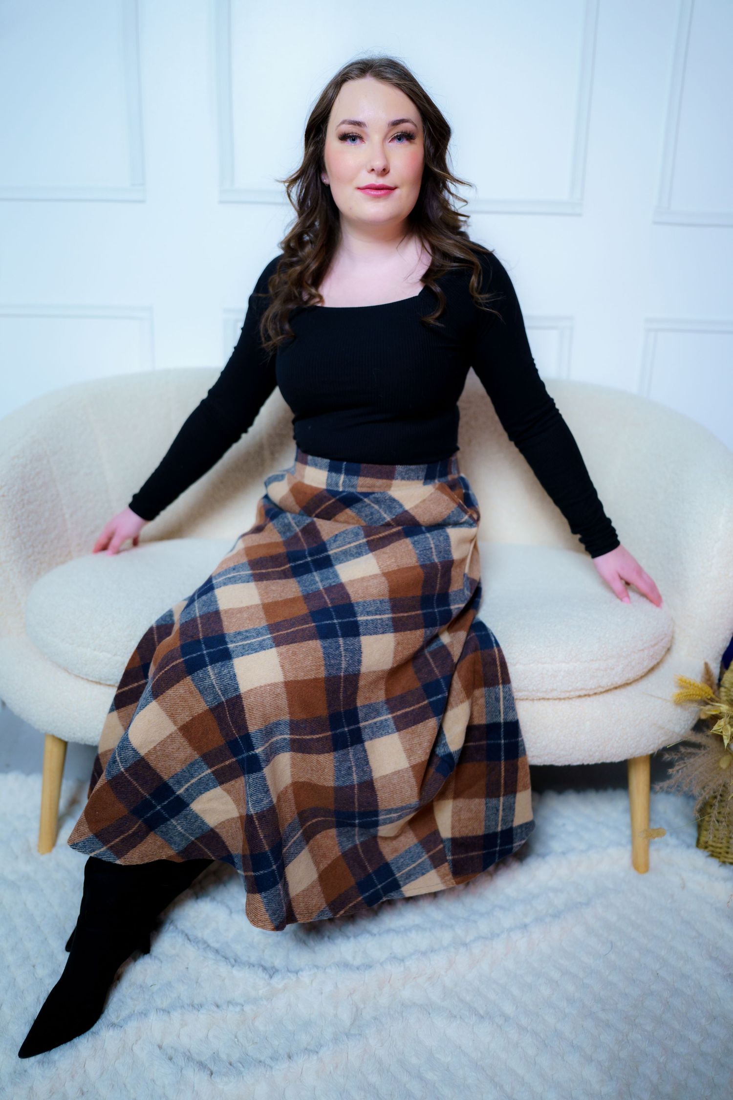 A person in a black top and plaid brown skirt sits elegantly on a cream-colored accent chair.