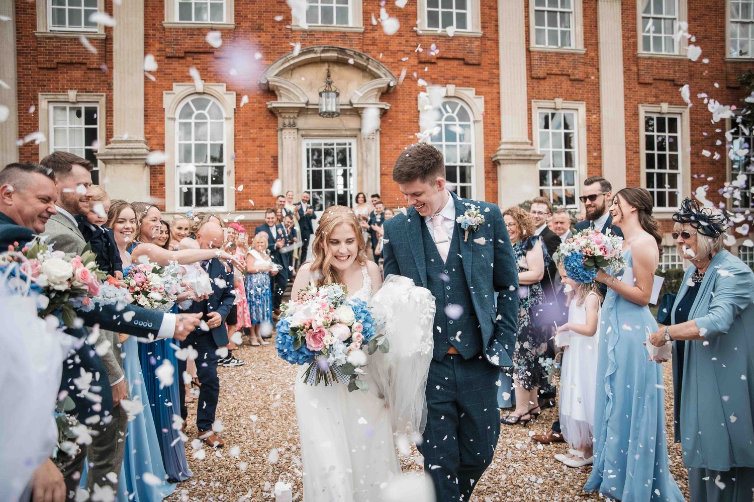 Wedding party celebrates with confetti outside a grand brick building with white windows as newlyweds walk past.