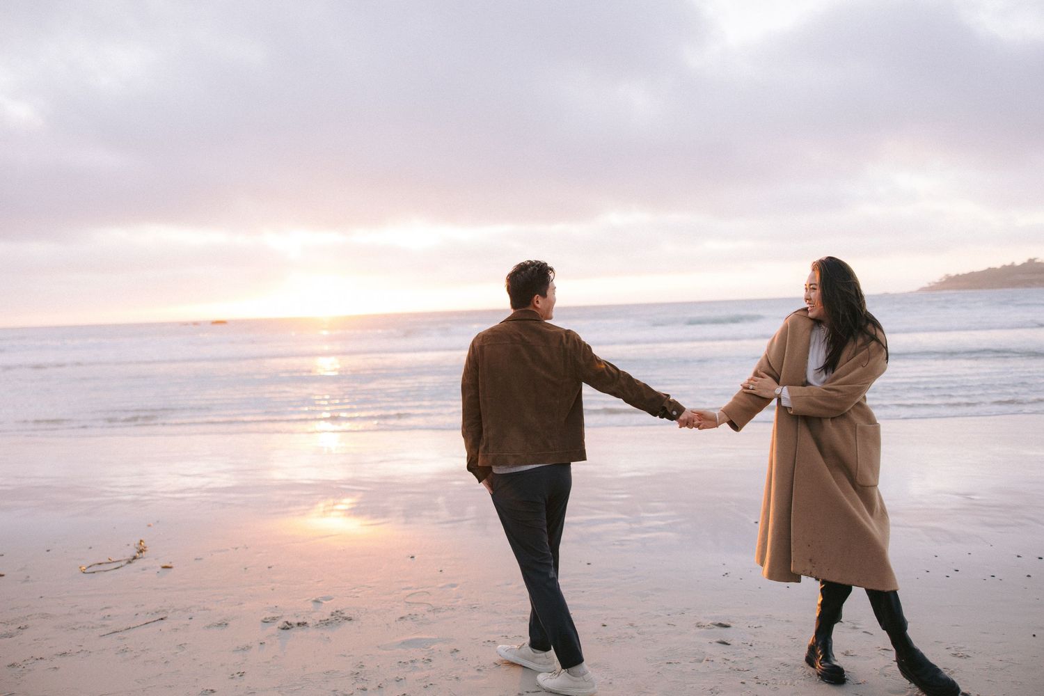 Two people holding hands while walking along a beach at sunset with waves crashing on the shore.