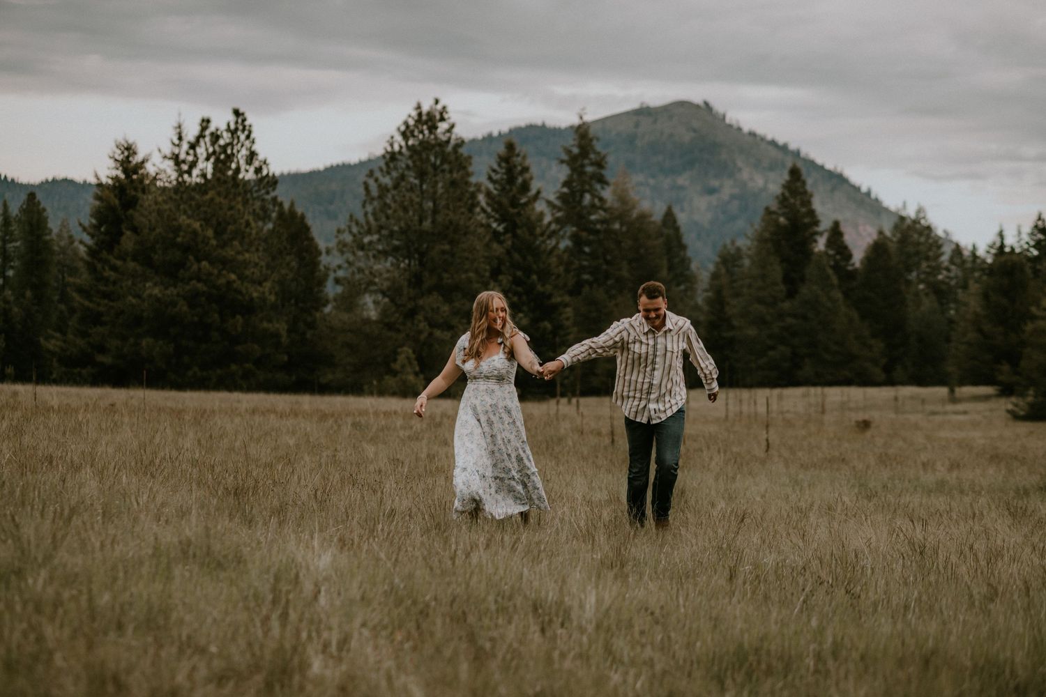 A couple holds hands and dances playfully together in a scenic mountain meadow at sunset.