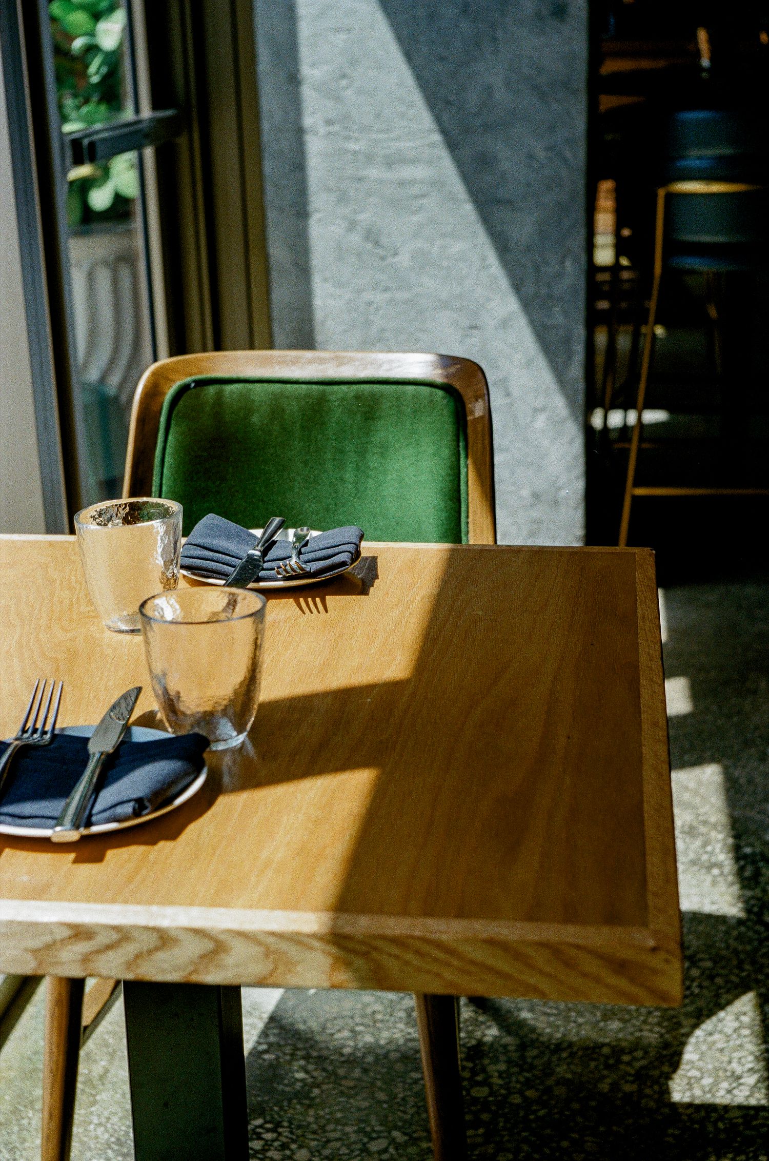 Place settings on a wooden restaurant table with green chairs and folded gray napkins in natural sunlight.