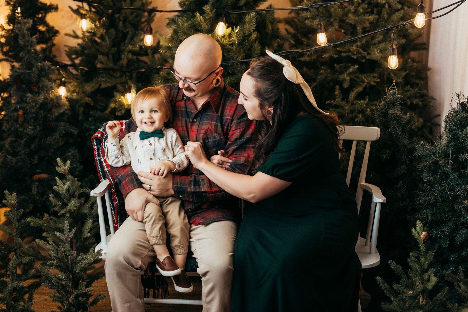 Family moment captured on a rocking chair surrounded by Christmas trees and warm lighting.