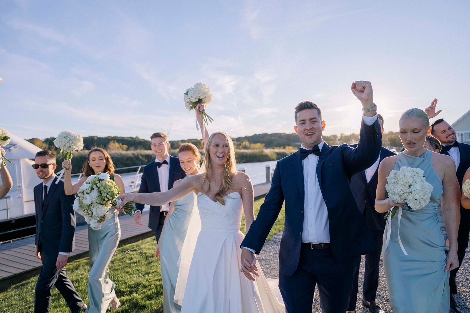Wedding party celebrates joyfully while walking along a scenic waterfront dock on a beautiful sunny day.